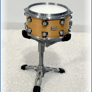 May include: A miniature drum set, featuring a tan drum with silver hardware and a white drumhead. The drum is mounted on a silver stand with a cross-shaped base and black feet. The drum set is a musical instrument.