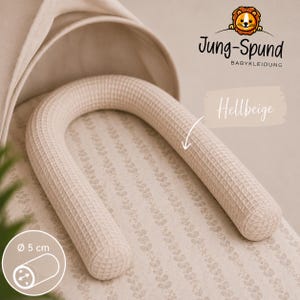 Könnte beinhalten: Ein hellbeiges, U-förmiges Waffel-Stillkissen auf einem gemusterten Stoffuntergrund. Das Kissen hat einen Durchmesser von 5 cm. Das Bild enthält den Text Jung-Spund BABY KLEIDUNG, das Wort Hellbeige und ein grafisches Symbol.
