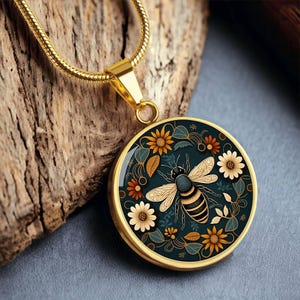 May include: Gold-toned pendant necklace featuring a detailed bee design surrounded by flowers and leaves. The round pendant has a dark teal background with a gold-colored frame. The necklace is displayed on a wooden surface.