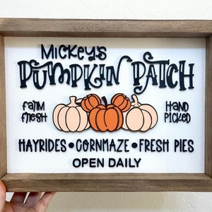 May include: A white wooden sign with a brown frame, advertising Mickey's Pumpkin Patch. The sign features the text "Mickey's Pumpkin Patch", "farm fresh", "hand picked", "hayrides", "corn maze", "fresh pies", and "open daily". The sign also features three pumpkins, one orange and two peach.