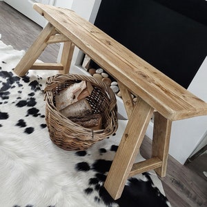 May include: A rustic wooden bench with a natural finish, featuring a wide, flat top and a simple A-frame design. The bench is placed on a white and black cowhide rug, with a wicker basket filled with firewood beside it.