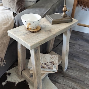 May include: A light-coloured, rustic wooden side table with a lower shelf. A white bowl with black cross designs sits on top, along with a small lamp and a stack of books. The table is placed on a cowhide rug.