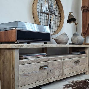 May include: A rustic wooden entertainment unit with two drawers and a shelf. The unit is made of reclaimed wood and has a natural finish. It is sitting on a cowhide rug in front of a white wall. A record player is sitting on top of the entertainment unit.