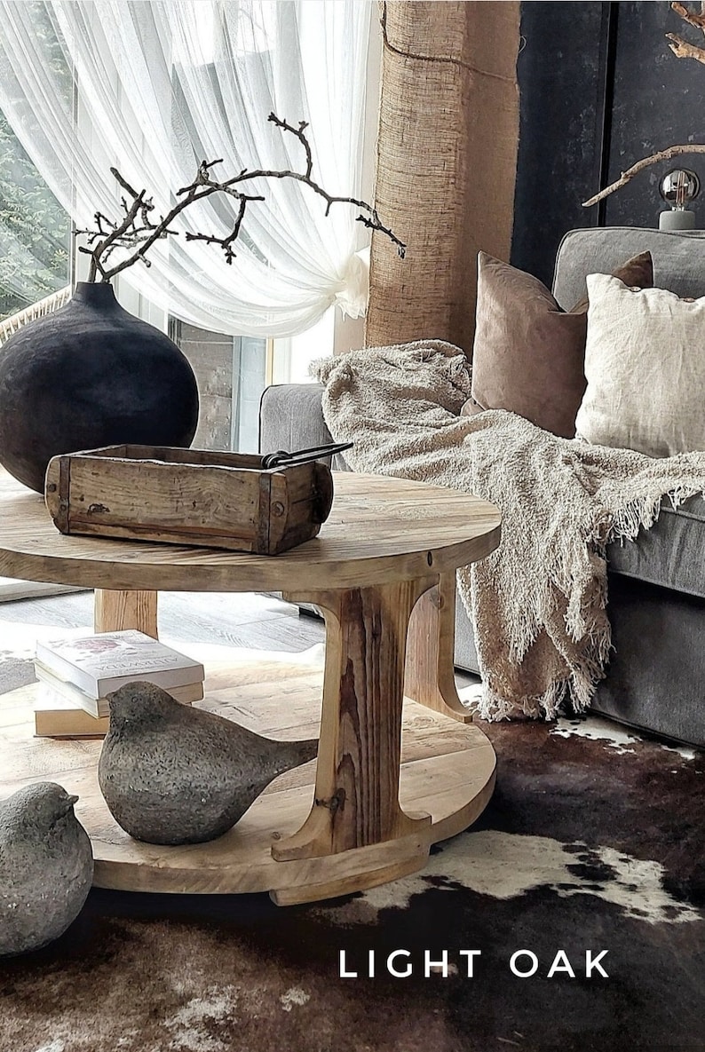 May include: A round wooden coffee table with a light oak finish. The table has a lower shelf and features a decorative wooden bird figurine. The table is sitting on a cowhide rug in front of a gray sofa with a white throw blanket.