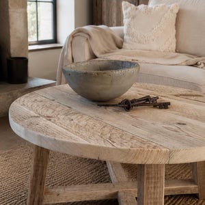 May include: A round wooden coffee table with a light brown finish and a cross-base design. A grey, weathered, circular bowl sits on the tabletop next to a small cluster of three dark, rusted metal skeleton keys resting on the wood surface.