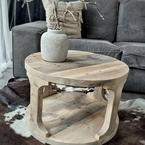 May include: A round wooden coffee table with a natural wood finish and a lower shelf. The table is 60 cm in diameter and has a rustic, reclaimed look. It is sitting on a cowhide rug in front of a gray sofa.