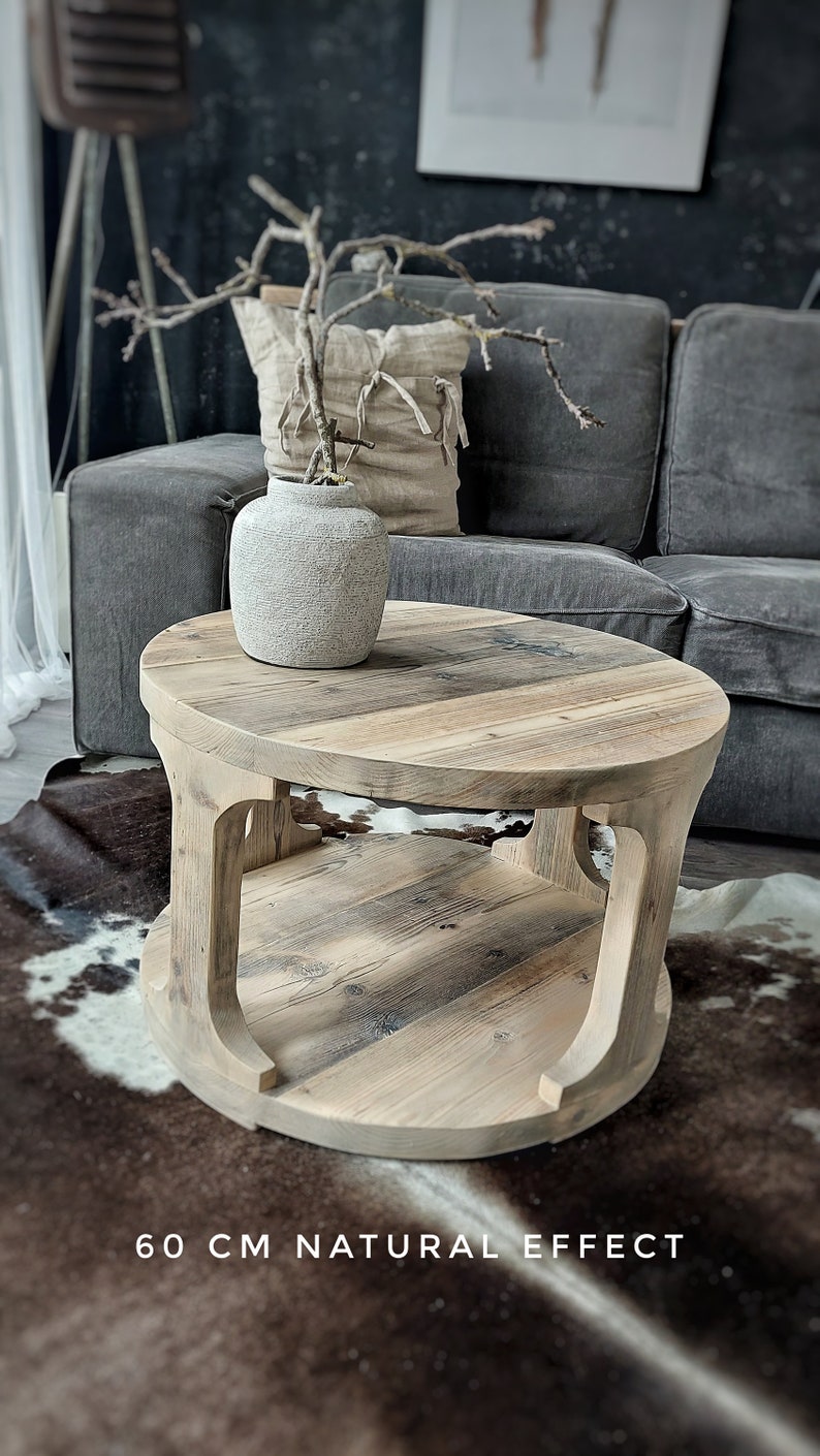May include: A round wooden coffee table with a natural wood finish and a lower shelf. The table is 60 cm in diameter and has a rustic, reclaimed look. It is sitting on a cowhide rug in front of a gray sofa.