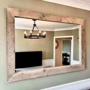 May include: A large rectangular mirror with a rustic wooden frame. The frame is made of reclaimed wood and has a natural, weathered finish. The mirror is reflecting a room with a chandelier and a television.