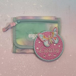 Könnte beinhalten: Ein kleines Portemonnaie mit Reißverschluss und einem pastellfarbenen Batikmuster. Auf der Vorderseite ist das Wort "ergo" aufgedruckt. Ein pinkfarbener, kreisförmiger Aufnäher mit dem Namen "Laura" und einem Schmetterlingsdesign liegt neben dem Portemonnaie.