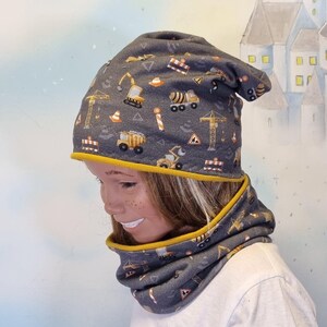 Może przedstawiać: Szary zestaw czapki i komina z motywem budowlanym. Tkanina przedstawia żółte, pomarańczowe i białe pojazdy budowlane oraz znaki bezpieczeństwa. Czapka i komin są obszyte żółtą tkaniną.