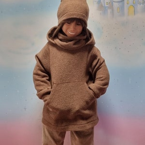 Könnte beinhalten: Ein braunes Kapuzenpullover mit einer großen Tasche auf der Vorderseite. Der Pullover wird von einem Kind getragen und hat eine braune Strickmütze auf dem Kopf. Das Kind steht auf einer rosa Oberfläche.