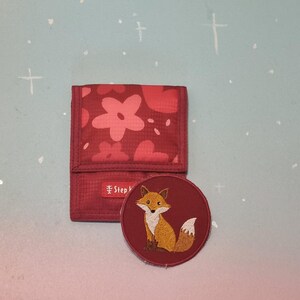 Op de afbeelding: Een rode portemonnee met een bloemenpatroon en een ronde patch met een geborduurde vos. De portemonnee heeft een rechthoekige vorm en een druksluiting. De vos patch staat op een bordeauxrode achtergrond.