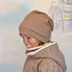 Puede incluir: Un gorro tejido en gofre de color beige y una bufanda con capucha a juego. El gorro tiene un borde doblado y un patrón texturizado. La bufanda tiene un ribete blanco y esponjoso alrededor de la capucha. El conjunto es de color neutro, adecuado para el frío.