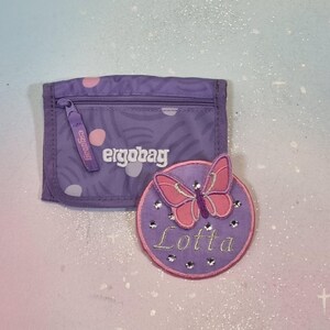 Könnte beinhalten: Ein lila Portemonnaie mit dem Markennamen "ergobag" und einem passenden runden Aufnäher. Der Aufnäher zeigt einen rosa Schmetterling und den Namen "Lotta" mit Strasssteinen.