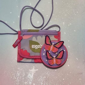 Puede incluir: Una pequeña cartera rosa y morada con un parche de mariposa a juego. La cartera tiene un estampado floral y la palabra "ergob" impresa. El parche de mariposa tiene dos mariposas con alas rosas y detalles morados.