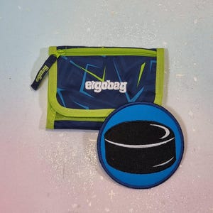 Op de afbeelding: Een blauw en groen portemonnee met de merknaam "ergobag" in het wit. De portemonnee heeft een geometrisch patroon en een bijpassende patch met een zwart hockeypuck-ontwerp op een blauwe achtergrond.