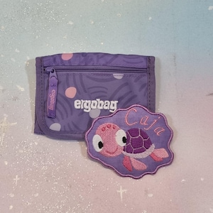 Könnte beinhalten: Ein lila Portemonnaie mit Reißverschluss und dem Markennamen "ergobag" aufgedruckt. Ein passender lila Aufnäher mit einem Cartoon-Schildkröten-Design und dem Namen "Caja" befindet sich neben dem Portemonnaie.