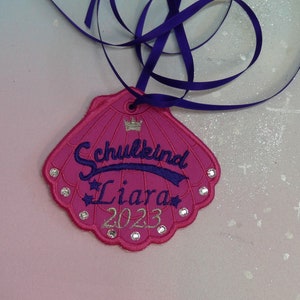 Op de afbeelding: Roze schelpvormig label met de tekst "Schulkind Liara 2023" geborduurd in paars en zilver. Het label heeft een paars lint.