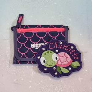 Può includere: Un portafoglio blu navy con cerniera rosa e motivo a scaglie rosa. Una toppa viola a forma di tartaruga con il nome "Charlotte" ricamato in rosa e un disegno di tartaruga con dettagli verdi e rosa.