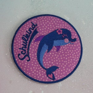 May include: A pink and blue embroidered patch with a dolphin and the word "Schulkind" in German.