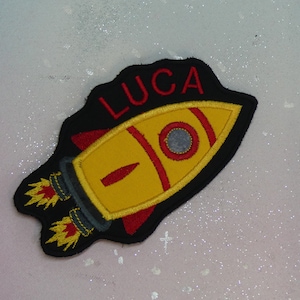 Può includere: Toppa a forma di razzo giallo con dettagli rossi e il nome "LUCA" in rosso. La toppa ha un bordo nero e fiamme. Adatta per personalizzare abbigliamento o accessori per bambini.