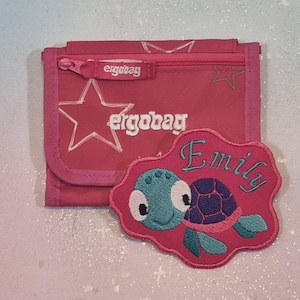 May include: A pink tri-fold wallet featuring the white text ergobag and star outlines. Attached to the front is a pink embroidered patch shaped like a cloud, displaying a teal and purple turtle graphic and the embroidered name Emily in teal.