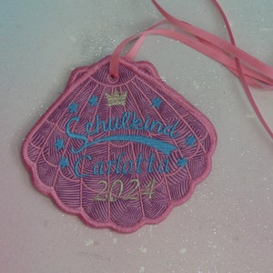 Könnte beinhalten: Rosa gestickter Patch in Muschelform mit einer Krone, dem Text "Schulkind Carlotta 2024" und blauen Sternen. Der Patch ist an einem rosa Band befestigt.