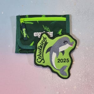 Puede incluir: Una cartera verde oscuro con cierre de cremallera y un parche bordado verde que muestra un delfín gris, el texto "Schulkind" en letra negra y el año "2025" en números negros. La cartera tiene un patrón de rayos.