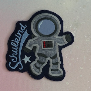 Puede incluir: Un parche bordado azul y gris de un astronauta con el texto "Schulkind" en una fuente azul cursiva.
