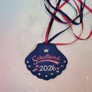 Op de afbeelding: Een donkerblauwe, schelpvormige hanger met geborduurde roze tekst Schulkind en het jaartal 2026 in het wit. Het ontwerp bevat een kleine kroon, roze sterren en heldere strass-steentjes langs de rand, opgehangen aan rode en blauwe linten.
