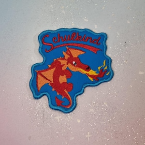 Puede incluir: Un parche bordado azul con un diseño de dragón rojo y naranja. El dragón está escupiendo fuego. La palabra "Schulkind" está escrita en rojo en la parte superior. El parche tiene un borde azul.