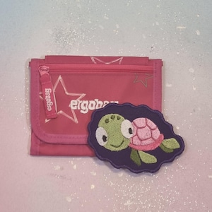 Könnte beinhalten: Ein pinkfarbenes Portemonnaie mit Reißverschluss und dem Markennamen "ergobag" auf dem Reißverschlussanhänger. Ein schildförmiger Aufnäher mit einer grünen Schildkrötenkopf und einem rosa Panzer liegt neben dem Portemonnaie. Das Portemonnaie hat ein Sterndesign.