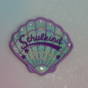 Puede incluir: Un parche en forma de concha con el texto "Schulkind 2024". El parche presenta una combinación de colores turquesa y morado, adornado con pedrería y detalles de estrellas. Una pequeña corona plateada en la parte superior.