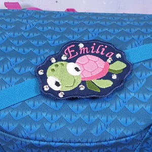 Pode incluir: Uma mochila azul com um padrão de escamas repetitivo apresenta um emblema bordado azul-escuro em forma de nuvem. O emblema exibe uma ilustração de uma tartaruga verde e rosa, o nome Emilia em letra cursiva rosa e vários strass decorativos transparentes.