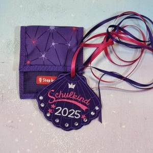 Op de afbeelding: Een paarse portemonnee met een geometrisch patroon en een bijpassend schelpvormig label. Het label is geborduurd met het woord "Schulkind" en het jaar "2025", plus decoratieve strass-steentjes en een kroon.