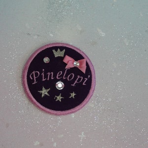 Könnte beinhalten: Ein kreisförmiger, gestickter Aufnäher mit dem Namen "Pinelopi" in Schreibschrift. Der Aufnäher ist dunkelviolett mit einem rosa Rand, einer kleinen rosa Schleife, einer silbernen Krone und Sternen. Strasssteine sind ebenfalls enthalten.