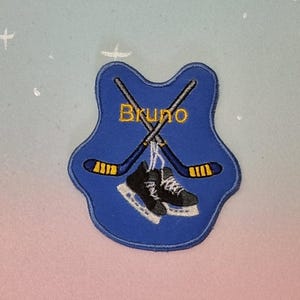 May include: A blue patch with a shield shape, featuring crossed hockey sticks and ice skates. The word "Bruno" is embroidered in gold. The hockey sticks have yellow and black details. The skates are black and white.