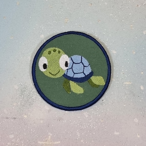 Op de afbeelding: Geborduurde patch met een cartoon schildpad. De schildpad heeft een groene kop en ledematen, een blauw schild en grote witte ogen. De patch is cirkelvormig, met een donkergroene achtergrond en een marineblauwe rand.