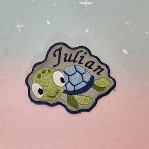 Op de afbeelding: Geborduurde patch met een cartoon schildpad met de naam "Julian". De schildpad heeft een groene kop en vinnen, een blauw schild en grote witte ogen. De patch staat op een grijze achtergrond met een blauwe rand.
