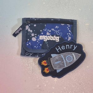 Könnte beinhalten: Ein rechteckiges blaues Portemonnaie mit weißem Spritzmuster und dem Text ergobag, dazu ein dunkelblauer Aufnäher mit dem gestickten Namen Henry und einer grauen Rakete mit orangefarbenen und gelben Flammen an den Triebwerken.