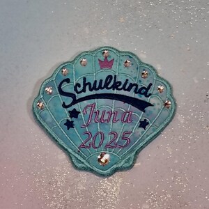 Op de afbeelding: Een turquoise schelpvormige patch met het Duitse woord "Schulkind" in marineblauwe letters, "Juna 2025" in roze letters en een kleine paarse kroon. De patch is versierd met strass-steentjes.