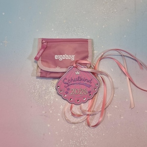 Op de afbeelding: Een roze portemonnee met de tekst ergobag en een bijpassende roze schelpvormige hanger met de tekst Schulkind 2026, een klein kroonicoontje en strass-steentjes. De hanger is bevestigd aan lange roze linten om op te hangen of te versieren.