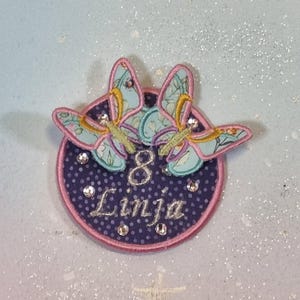 Puede incluir: Un parche bordado redondo con dos mariposas con alas azul claro y diseños florales. El parche tiene un fondo azul oscuro con lunares blancos, un borde rosa y el número "8" y el nombre "Linja" bordados en plata.