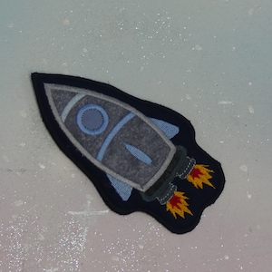 Könnte beinhalten: Eine blaue und graue Filz-Raketen-Applikation mit gelben Flammen. Die Rakete befindet sich auf einem weißen Hintergrund mit einem Glitzer-Effekt.