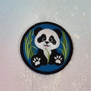 Könnte beinhalten: Runder, gestickter Aufnäher mit einem Panda-Bären mit schwarz-weißem Fell. Der Panda sitzt mit ausgestreckten Pfoten, umgeben von grünen Bambusstängeln auf blauem Grund, mit einem schwarzen Rand.