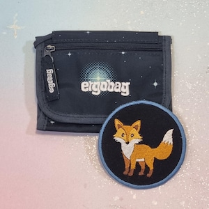 May include: A dark blue wallet featuring a starry night pattern and the white text ergobag on the front, accompanied by a circular black patch with an embroidered orange and white fox design, featuring a blue border around the edge of the patch.