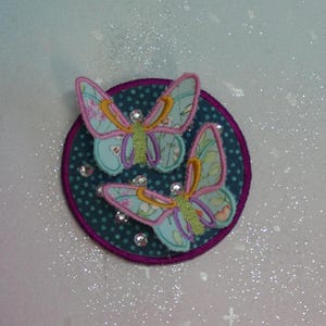 Peut inclure: Deux papillons bleus et roses avec des accents jaunes sont cousus sur un cercle de tissu turquoise à pois blancs et une bordure violette.