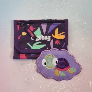 Op de afbeelding: Een paarse portemonnee met een kleurrijk abstract patroon en de merknaam "ergobag". Een bijpassende paarse patch met een lachende cartoon schildpad ligt naast de portemonnee. De schildpad heeft een groene kop, een paarse schild en gele vinnen.