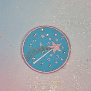 Pode incluir: Um patch redondo bordado com fundo azul claro e borda rosa. O patch apresenta uma estrela rosa, estrelas rosas menores e um design de estrela cadente com detalhes dourados e brancos. Strass transparentes são espalhados pelo patch.