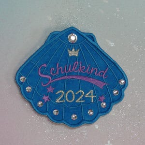 Könnte beinhalten: Blaue Filzmuschel mit Strasssteinen, einem lila Band mit dem Text "Schulkind 2024" und einer silbernen Krone.